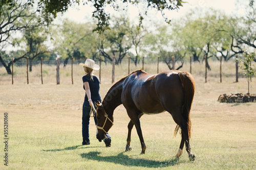 Western lifestyle on farm or ranch shows cowgirl walking with horse through rural field.
