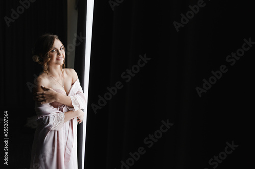 Smiling woman in the pink peignoir standing near the window. Morning of the bride. Bride portrait photo