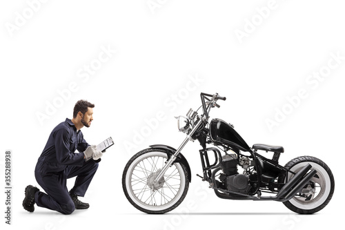 Fototapeta Motorbike mechanic in a uniform kneeling and looking at a chopper and writing a