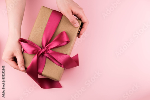 Female hands hold a gift box wrapped in craft paper and tied with a pink ribbon on a pink background close-up. Copy space © leks3003