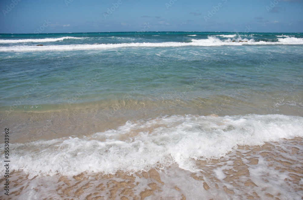 Peaceful blue water and ocean waves roll in on a shallow beach with taupe-colored sand and frothy white residue.. Lots of room for vacation dialog text.
