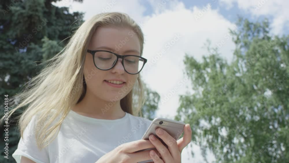 Joyful bespectacled girl with blonde hair smiles holding mobile phone in hands and answering to message on sunny summer day. Joyful bespectacled girl with blonde hair smiles holding mobile phone in hands and answering to message on sunny summer day.