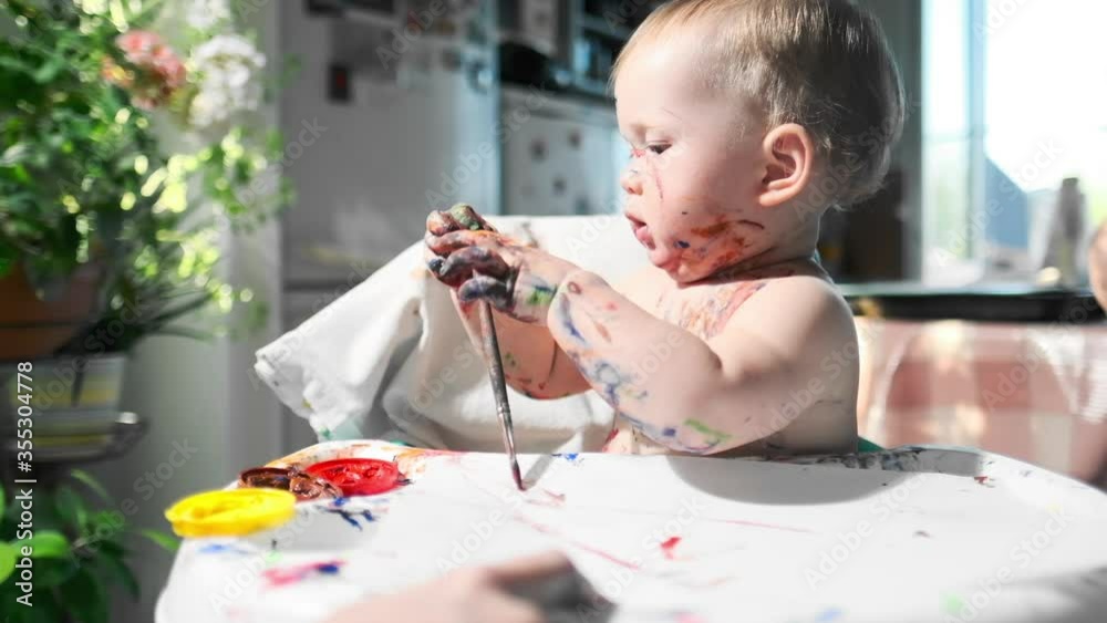 Baby paints at home. Portrait of the infant baby enthusiastically drawing with brushes and paints and coloring herself at home Baby paints at home. Portrait of the infant baby enthusiastically drawing with brushes and paints and coloring herself at home