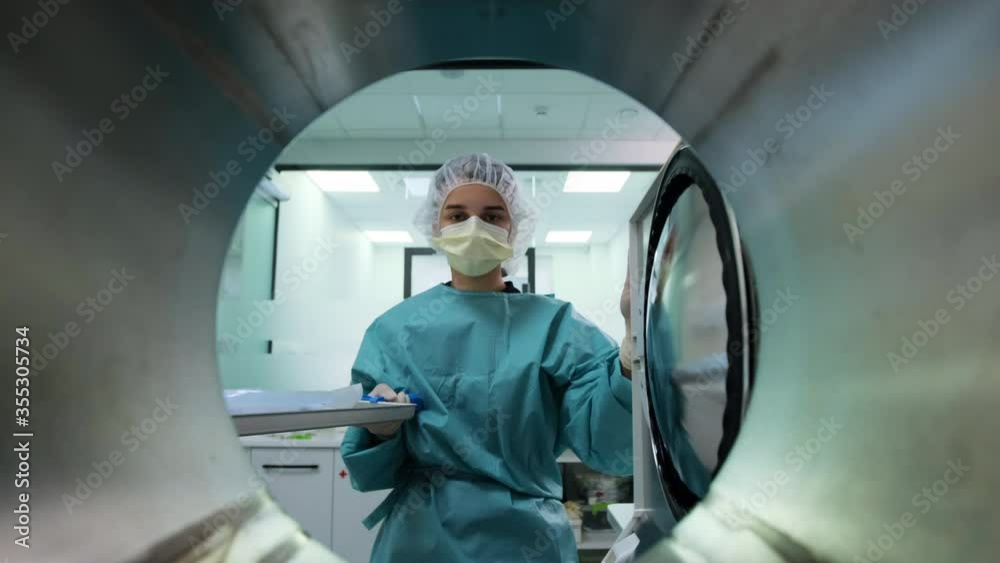 Laboratory assistant in green uniform, yellow mask and white cap takes out packaging with dental tools from autoclave sterilizer machine after disinfection. POV medical oven. Modern professional