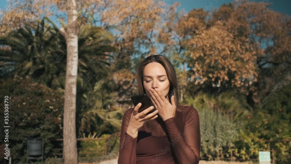 Tracking out shot of beautiful blond girl happily recording voice message and sincerely sending air kiss to her boyfriend by the phone outdoor. Attractive girl using mobile messenger Tracking out shot of beautiful blond girl happily recording voice message and sincerely sending air kiss to her boyfriend by the phone outdoor. Attractive girl using mobile messenger