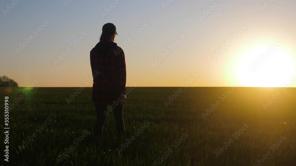 Farmer girl walking beside green wheat field enjoying the sun. Female farmer in plaid shirt and hat walks through the field. A worker go with his rubber boots at sunset time. Concept of agricultural. Farmer girl walking beside green wheat field enjoying the sun. Female farmer in plaid shirt and hat walks through the field. A worker go with his rubber boots at sunset time. Concept of agricultural.