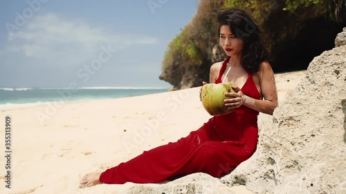 Woman quench her thirst with coco nut. Beauty, baunty, travel, fashion concept. Vacation on exotic islandBeautiful pretty asian woman in hat and red dress drinking coconut on tropical paradise beach. photo