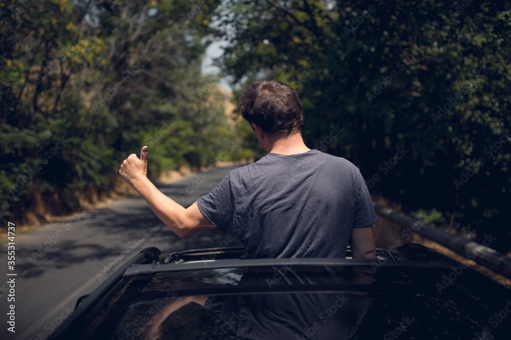 Young happy man drives a car and holds his hand out from the window. Driver enjoys driving and shows Ok sign with his hand out of window. Road trip, travel and freedom concept.