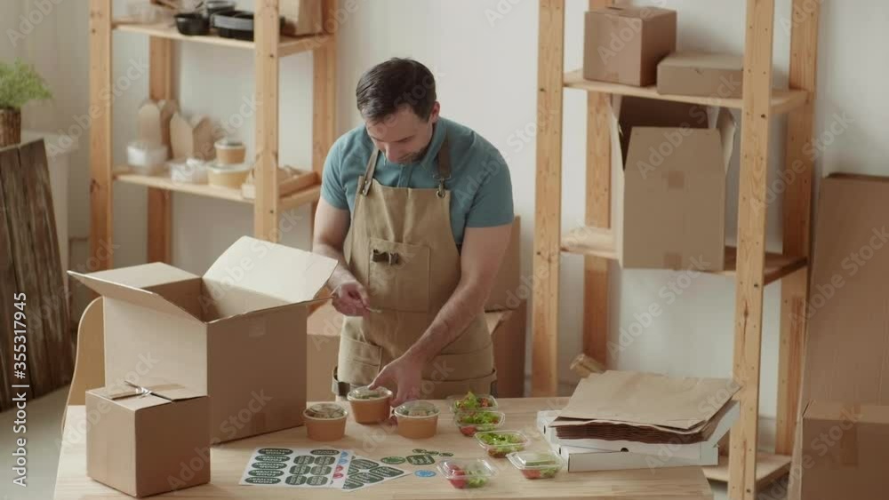 Medium shot of Caucasian man wearing apron standing at wooden table in office and packing box for delivery