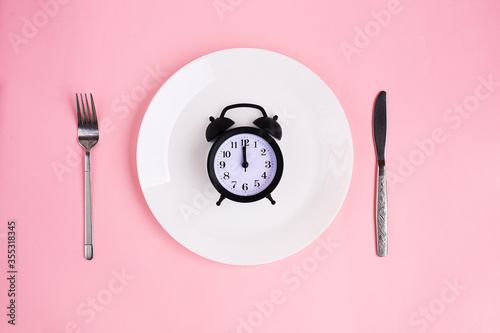 Time to eat. Lunch time. View from above on white plate with alarm clock and knife and pork near on pink background. 12 a.m.