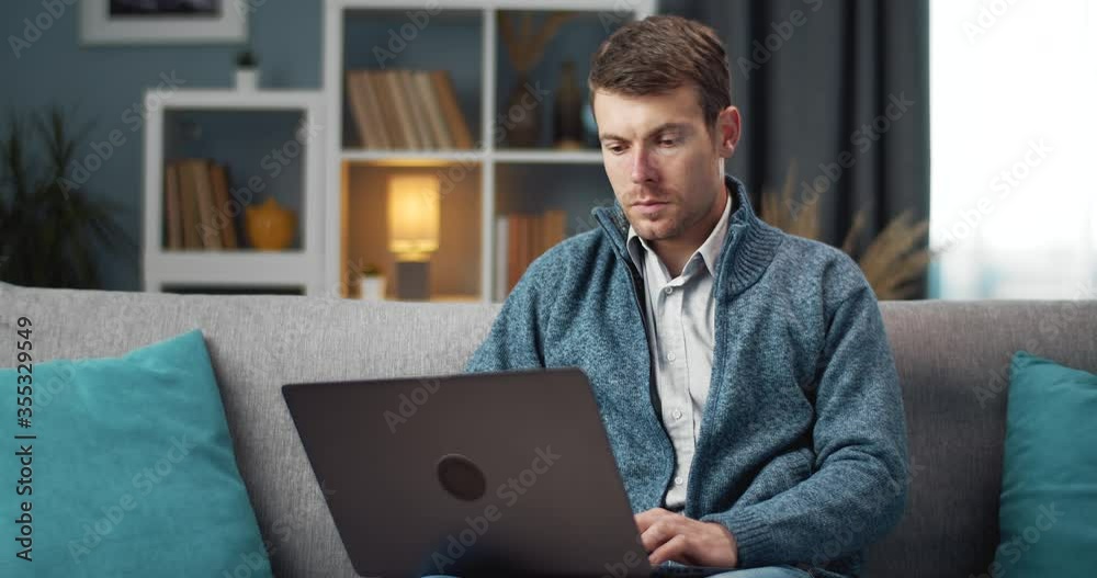 Bearded worker in domestic outfit using personal laptop while sitting on grey couch. Young guy spending free time at home for remote work. Bearded worker in domestic outfit using personal laptop while sitting on grey couch. Young guy spending free time at home for remote work.
