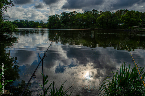 sun, clouds, and blue sky reflecting off a tranquil lake in waning light, fishing poles with lines in the water silhoetted in the foreground, ready for a peaceful night of catfishing  photo