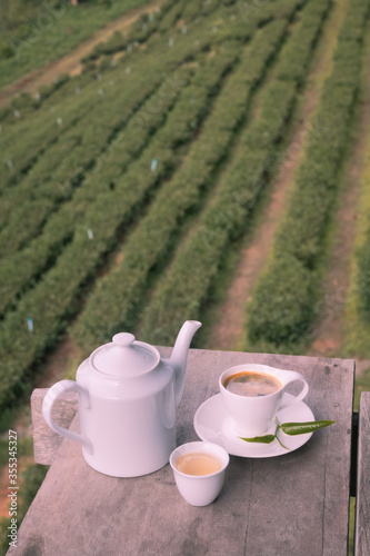 White ceramic teapot, teacup, and cup of fresh coffee on wooden table with Chinese tea plantation background