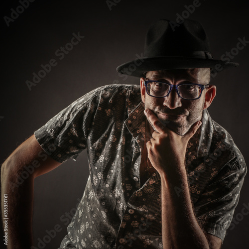 Portrait of a bald man with glasses, with stubble and a hat of a man who is sitting on a chair in a dark space. photo