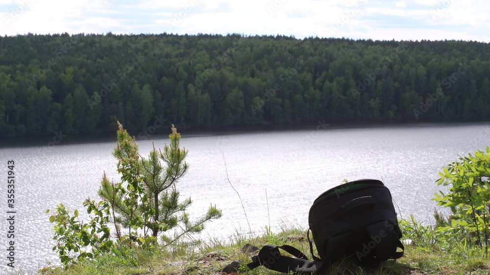 Obraz premium A moment from a trip by a mountain lake with a black backpack left on a cliff of a cliff against the background of a smooth surface of water and a dark forest on the other side