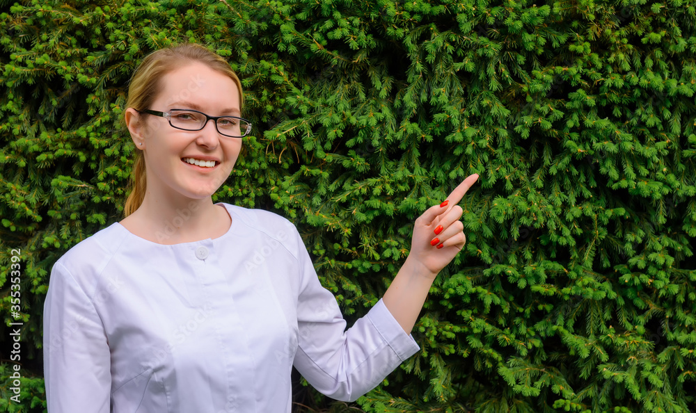 Female doctor with finger point up. Nutritionist in white coat and glasses on green foliage background with copy space. Image for advertising scientific developments in the food and medical industry. Female doctor with finger point up. Nutritionist in white coat and glasses on green foliage background with copy space. Image for advertising scientific developments in the food and medical industry.