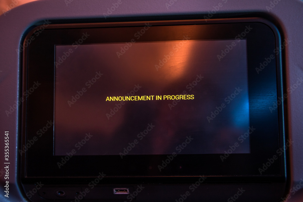 A monitor on backside of chair at airplane with text - announcement in progress - on display. Notice on screen in a flying aircraft. A monitor on backside of chair at airplane with text - announcement in progress - on display. Notice on screen in a flying aircraft.