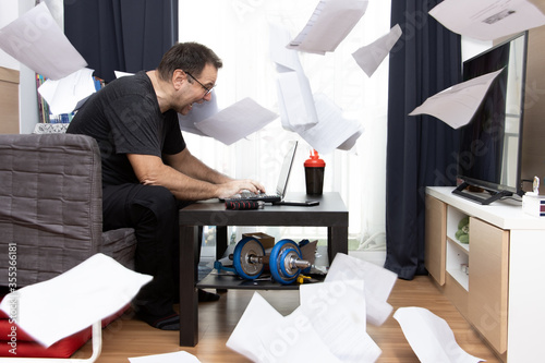 A dynamic zeal man sitting in living room and works on laptop, paper documents flying around. A Emotional Home office.