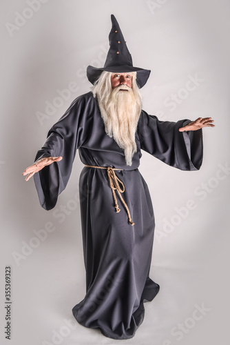 A stern grey-haired bearded wizard in a gray cassock and a cap is practicing sorcery and doing magic against a light background.