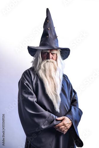 A severe grey-haired bearded sorcerer in a gray cassock and a cap is practicing sorcery and doing magic against a blue background.