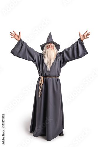 A stern grey-haired bearded wizard in a gray cassock and a cap is practicing sorcery and doing magic against a light background.