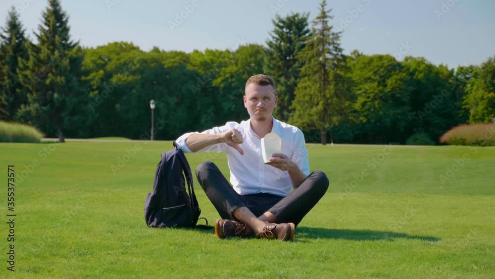 Businessman eats vegetarian lunch with an ecologic package, frowning and showing dislike. Guy sits in the park on the green grass eats a salad. Wooden sticks. Paper packaging Businessman eats vegetarian lunch with an ecologic package, frowning and showing dislike. Guy sits in the park on the green grass eats a salad. Wooden sticks. Paper packaging