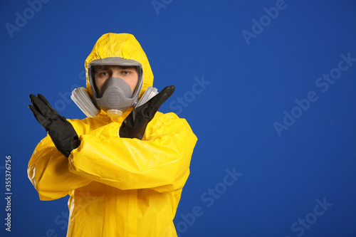 Man in chemical protective suit making stop gesture on blue background, space for text. Virus research