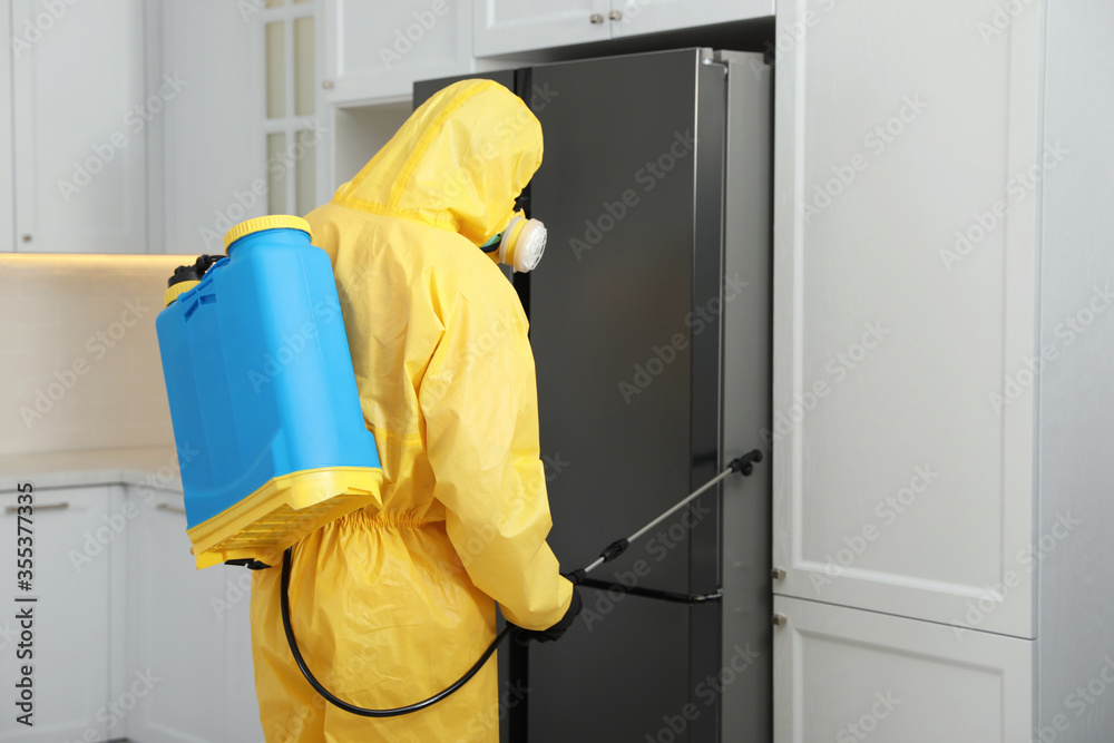 Pest control worker in protective suit spraying insecticide near refrigerator indoors Pest control worker in protective suit spraying insecticide near refrigerator indoors