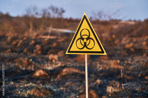 Horizontal snapshot of yellow biohazard sign installed in scorched field with burnt anthills on a blurred background. Concept of ecology and burned earth.