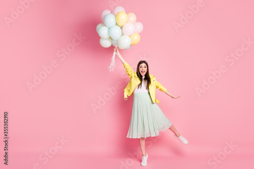 Full size photo of funny excited lady hold hand many air balloons flying up air wind blowing wear yellow leather jacket green long skirt shoes isolated pastel pink color background