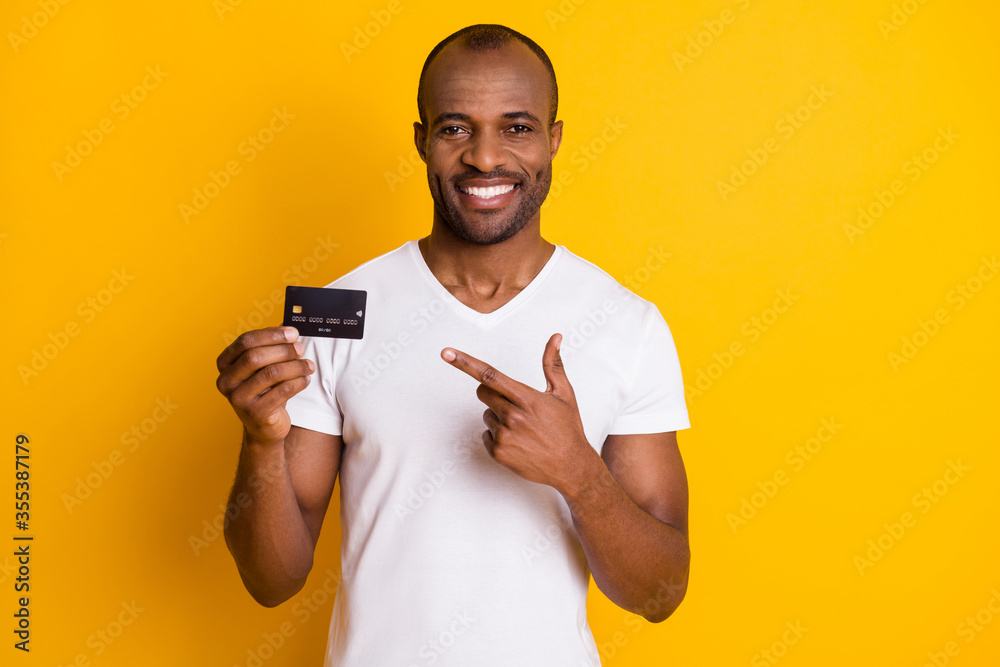 Portrait of positive afro american guy hold credit card suggest easy banking profit payment point index finger wear white t-shirt isolated over bright shine color background Portrait of positive afro american guy hold credit card suggest easy banking profit payment point index finger wear white t-shirt isolated over bright shine color background