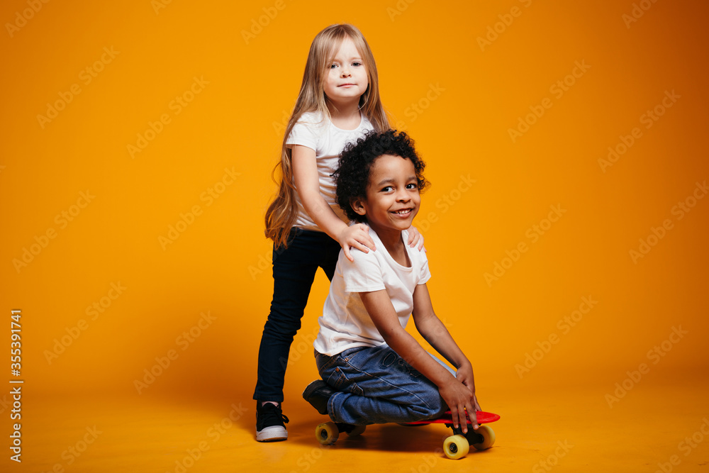 A curly boy sits on a skateboard, and behind him is a long-haired blonde girl and rolls the boy pushing him in the back. A curly boy sits on a skateboard, and behind him is a long-haired blonde girl and rolls the boy pushing him in the back.
