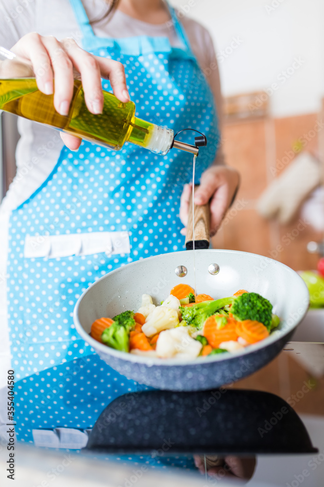Close up of housewife holding bottle of olive oil and cooking pan with various vegetables in kitchen.