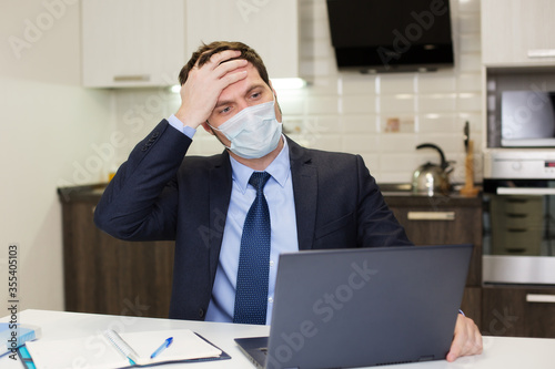 man in a mask holds his head with his hand and looks at the computer, work at home
