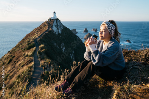 Beautiful asian tourist chilling out at Nugget Point, Dunedin, New Zealand. Young asian traveller enjoys coffee in morning at coastline of New Zealand. Lifestyle image of people in natural landscape.
