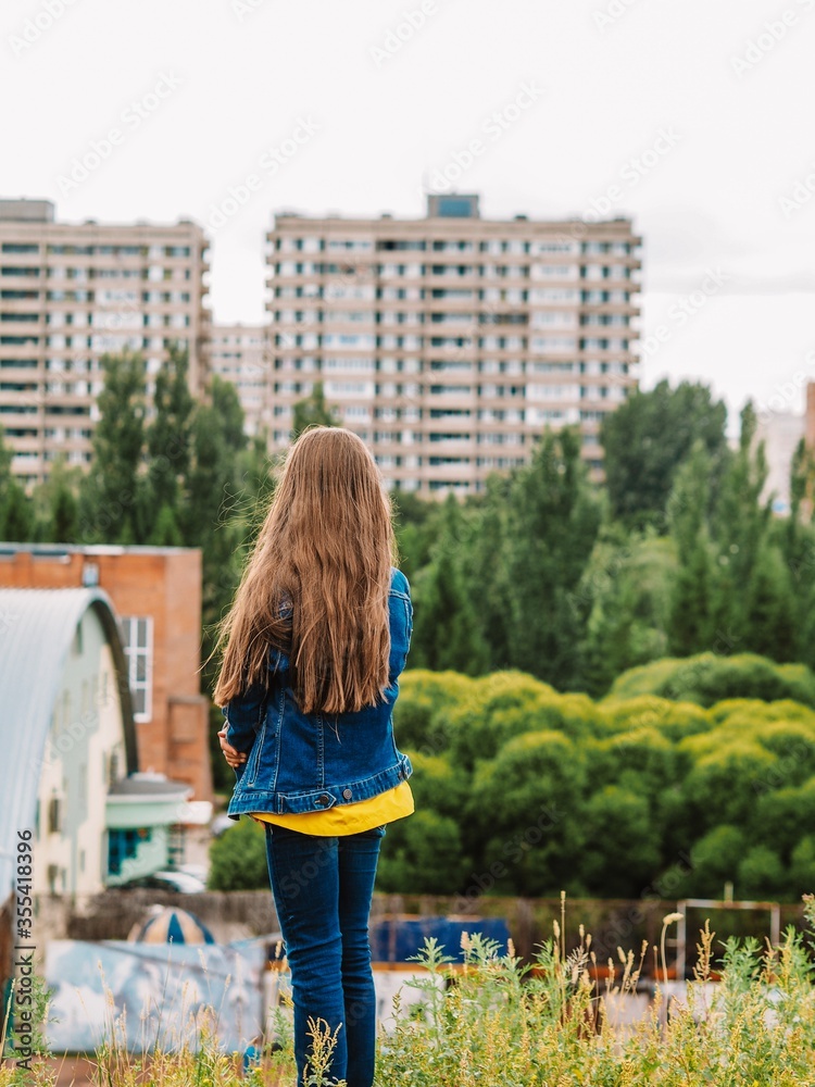 A teenage girl in a denim jacket with long hair turned away from the camera and looks down from a high mountain at the panorama of the city