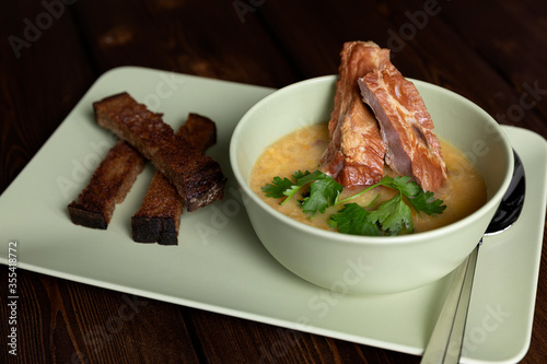 Homemade thick pea soup with smoked pork ribs in a deep ceramic bowl on a dark wooden table. A sprig of greenery. Rye bread croutons on a platter. Soup spoon.