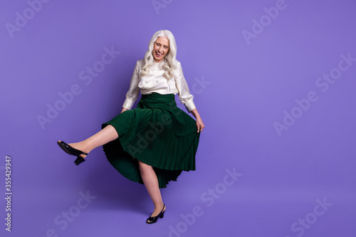 Full length photo of attractive aged white haired lady good mood hold long green skirt dancing pensioner retro party raise leg wear white blouse isolated pastel purple color background