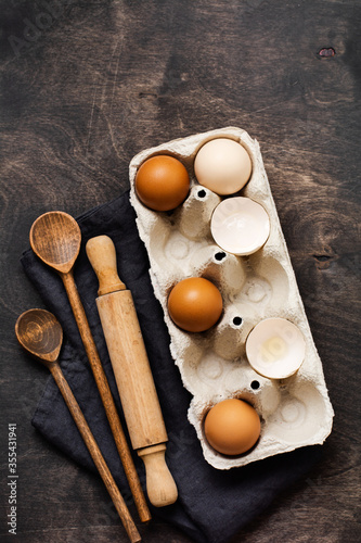 White and yellow chicken eggs and shells in an old cellulose vintage container with flour, sugar, wooden spoons and a rolling pin on old dark wooden background. Baking and culinary background. 