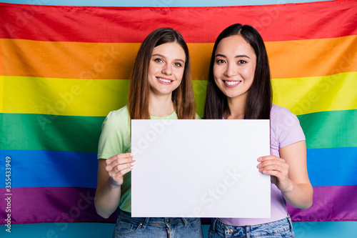 Photo of lesbians couple ladies protest parade day tolerance same sex marriages stand in front gay rainbow flag background hold billboards protesters wear casual t-shirts jeans