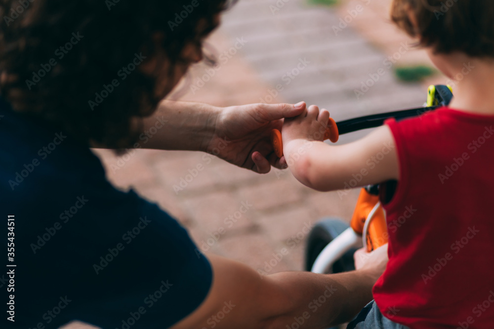 Father's Day. Dad with his little son on a bike from back. Family walk with a child. Outdoor activities for family. Dad helps son with a bike. Hands of father and small child.