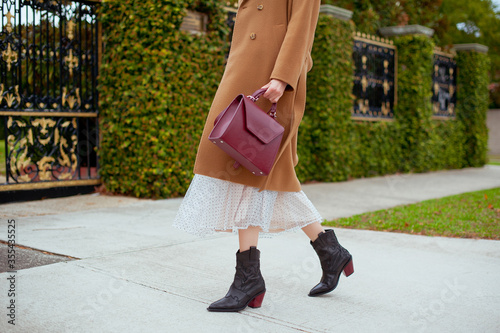 Detail of young fashionable woman wearing beige wool coat, tulle midi skirt and black high heel cowboy boots. She is holding stylish burgundy handbag in hands. Street style.
