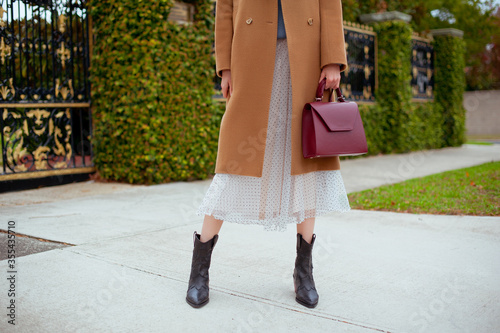 Detail of young fashionable woman wearing beige wool coat, tulle midi skirt and black high heel cowboy boots. She is holding stylish burgundy handbag in hands. Street style.