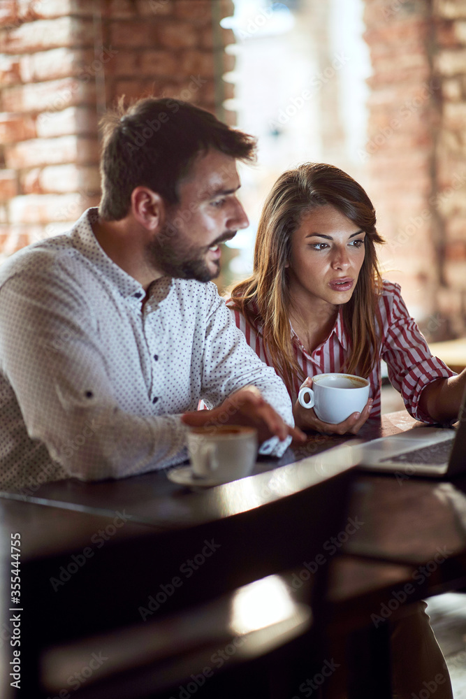 caucasian couple looking at laptop in front of them, illuminated by light of a monitor, talking, drinking coffee in cafe
