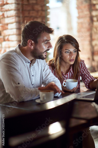 caucasian couple looking at laptop in front of them  illuminated by light of a monitor  talking  drinking coffee in cafe