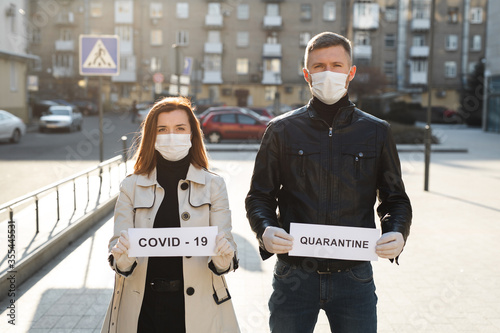 A woman and a man in protective medical masks hold a placard with the words Quarantine and Covid 19. © Artem Zakharov
