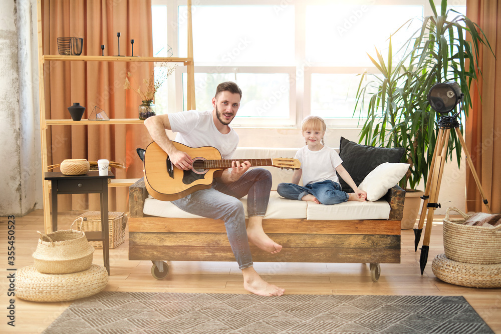 Handsome young man at home playing the guitar for his little cute girl. Happy Father's Day! Father and daughter in white t-shirts and jeans sit on couch in the living room. I love you, dad! Family Handsome young man at home playing the guitar for his little cute girl. Happy Father's Day! Father and daughter in white t-shirts and jeans sit on couch in the living room. I love you, dad! Family