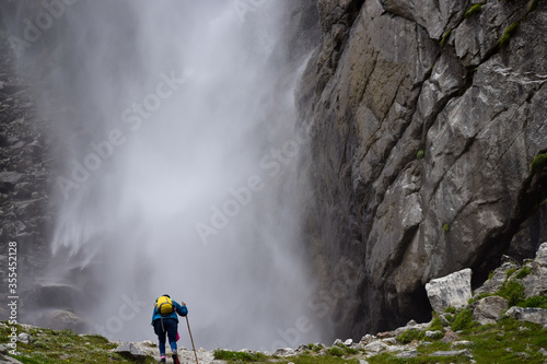 A solo trekker is approaching to the roaring Vasudhara falls surrounded by rocky brown mountains & clouds situated near Mana (last indian village). Monsoon trek taken in August in Uttarakhand India.