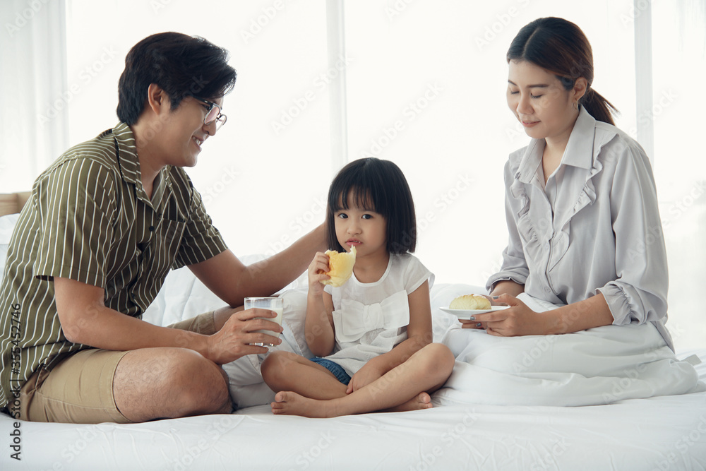 Happy Asian family having breakfast with milk bread sausages at bedroom, a little daughter eating sausage bread on bed at home Happy Asian family having breakfast with milk bread sausages at bedroom, a little daughter eating sausage bread on bed at home