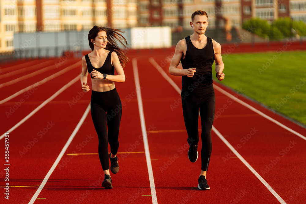 Young man and woman running wearing black sport suits in modern stadium. 